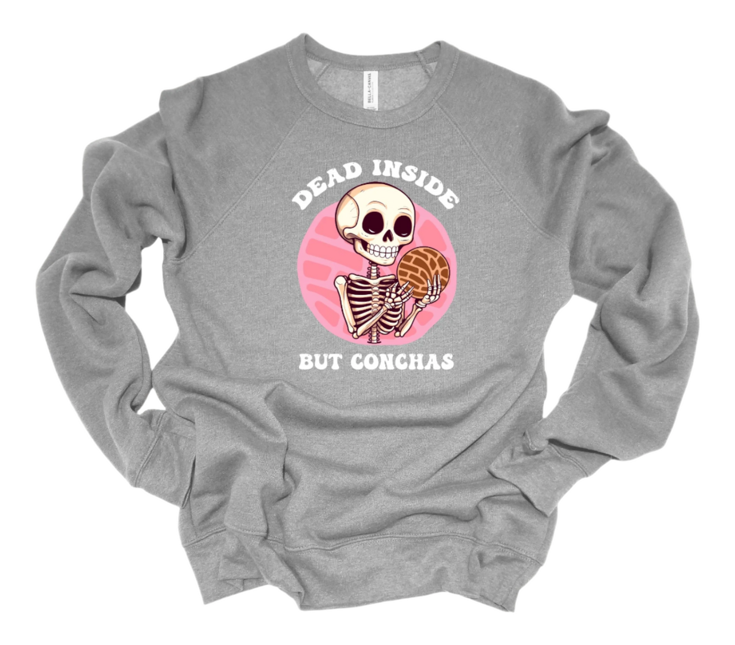A gray sweatshirt with a whimsical illustration of a spooky cute skeleton enjoying Mexican conchas, surrounded by the phrase 'Dead Inside but Conchas' in playful typography