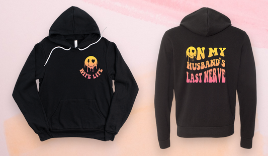 A black sweatshirt featuring a melty smiling face with a sunset gradient, accompanied by the text 'wife life' on the front and 'On my husband's last nerve' on the back
