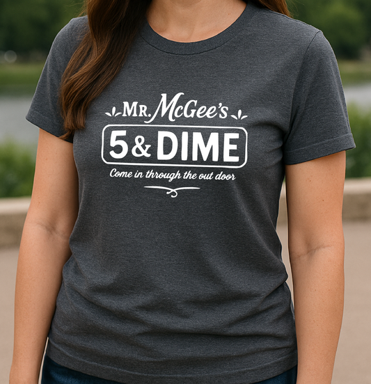 Woman wearing a dark gray t-shirt with 'Mr. McGee's 5 & Dime' text, standing outdoors with a cityscape in the background.